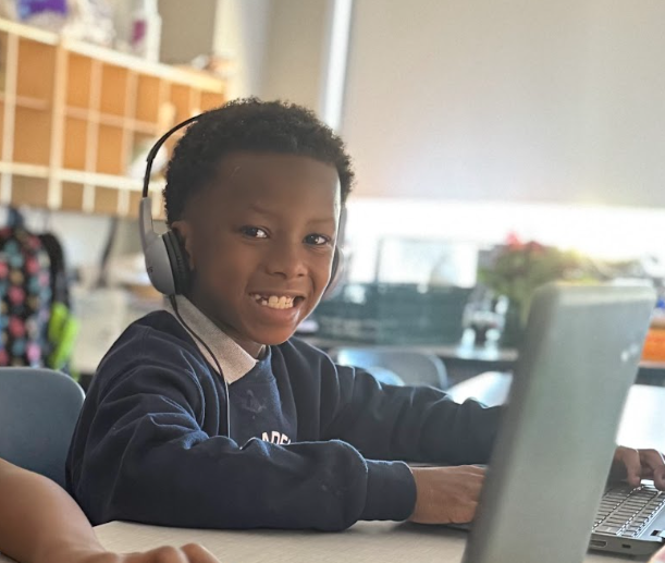 Image of a smiling elementary student sitting in a classroom at a table with a laptop. He is wearing a dark blue crewneck sweatshirt and a pair of headphones. There are cubbies in the background and another student's laptop in the foreground.