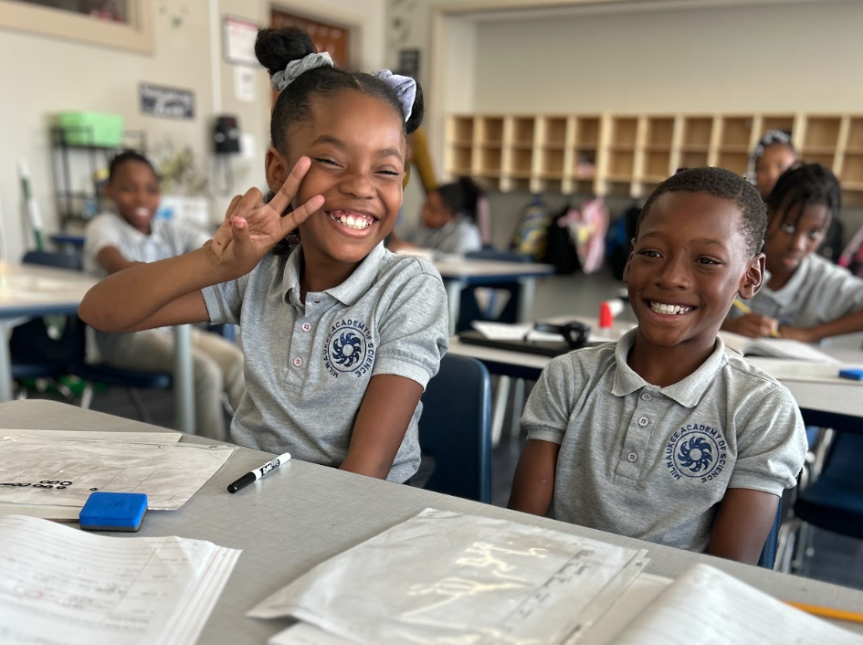 Students in a classroom, with two smiling students in the forefront, both in grey polo uniforms with the MAS logo on the right breast. The female student on the left is raising a peace sign to her face.