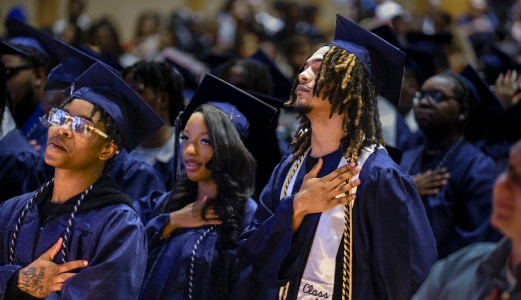 Focus on three high school seniors in a crowd of graduating students, standing with their hands over their hearts.