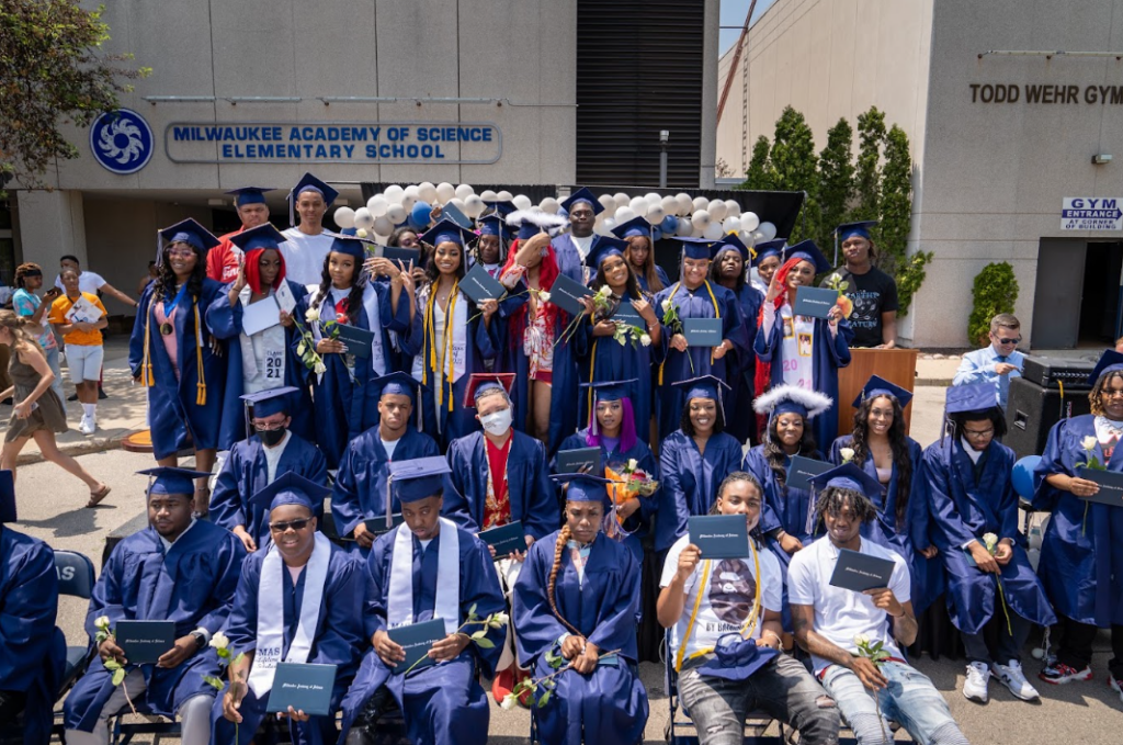 Graduating high school seniors wearing blue caps and gown in front of Milwaukee Academy of Science's elementary school entrance.