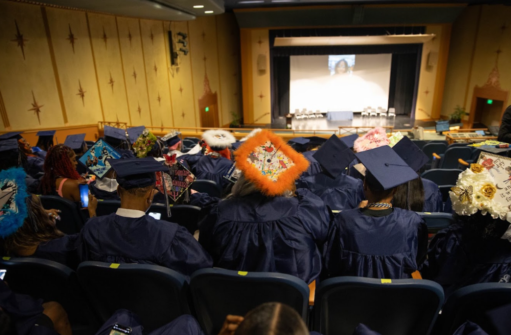 Back shot of graduating high school seniors wearing blue caps and gowns sit in an auditorium.