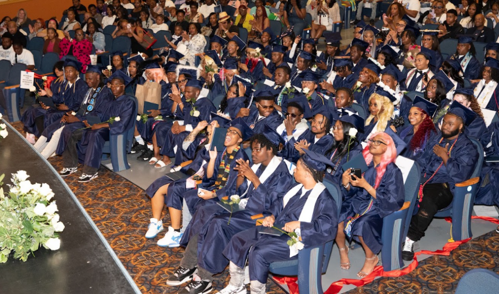 Graduating high school seniors in blue caps and gowns sit in an auditorium, surrounded by friends and family