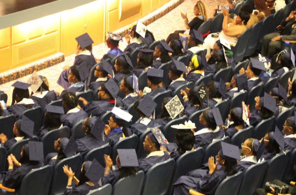 Back shot of graduating high school seniors wearing navy caps and gowns and sitting in an auditorium